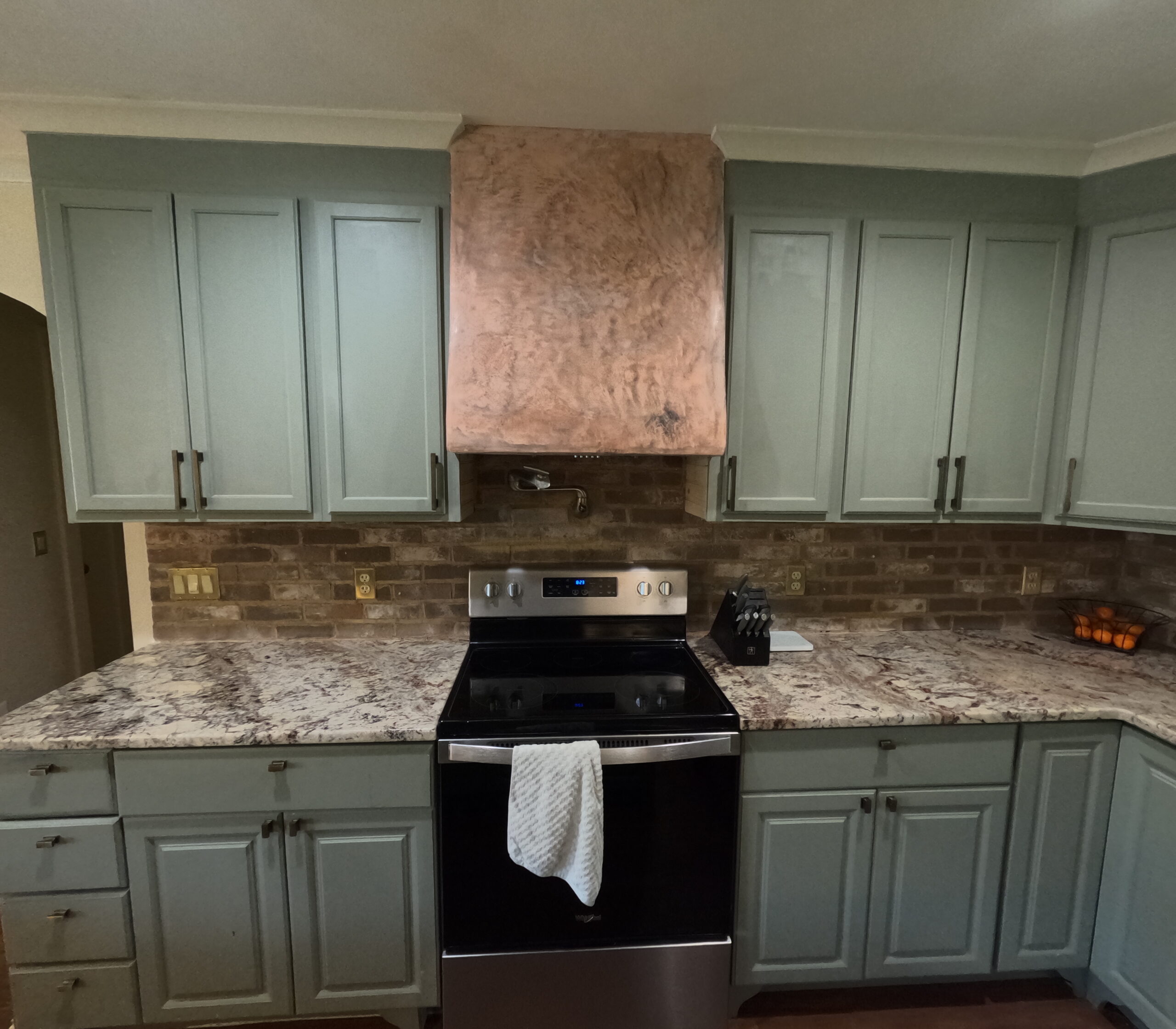 Sage kitchen cabinets with bronze hardware and trim feet either side of a tadelakt range hood